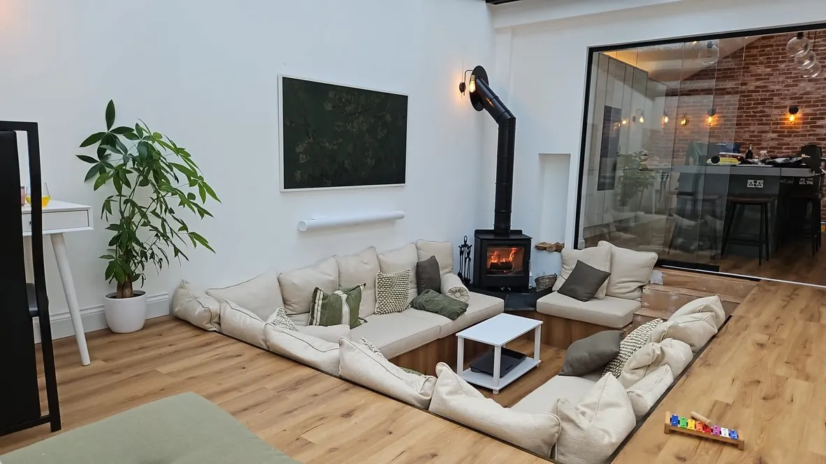 Furnished sunken lounge around a lit wood burner, kitchen visible through a glass screen